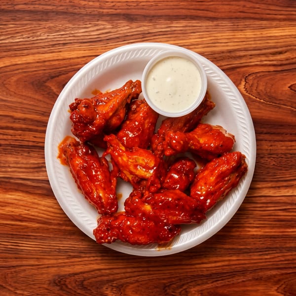 A white plate of red, saucy chicken wings with a side of ranch dressing on a wooden table.